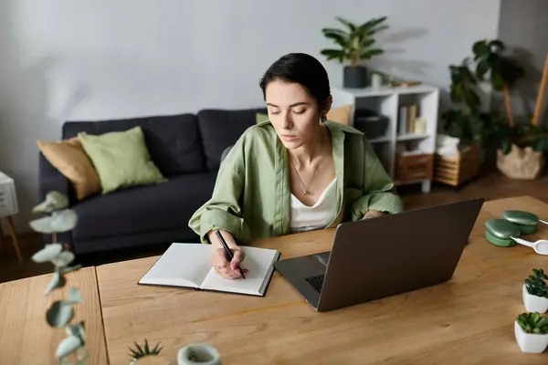 A woman sits at a desk in her home, writing in a notebook while her laptop is open in front of her. — Stock Photo