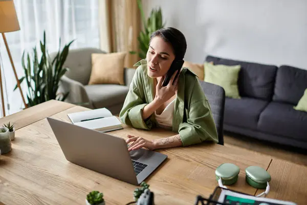 A woman works on her laptop and answers a phone call while sitting at a desk in a home office. — Stock Photo