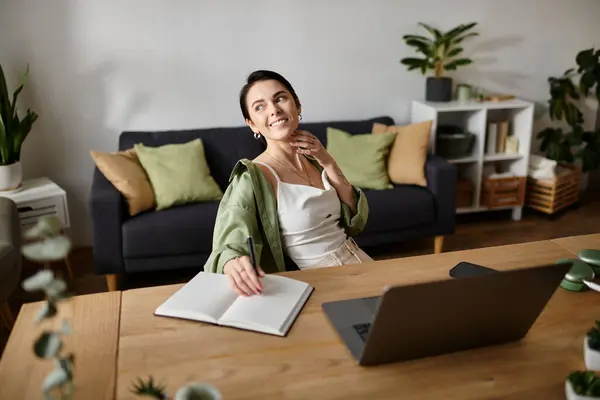 A woman sits at a desk in her home office, writing in a notebook while a laptop sits beside her. — Stock Photo