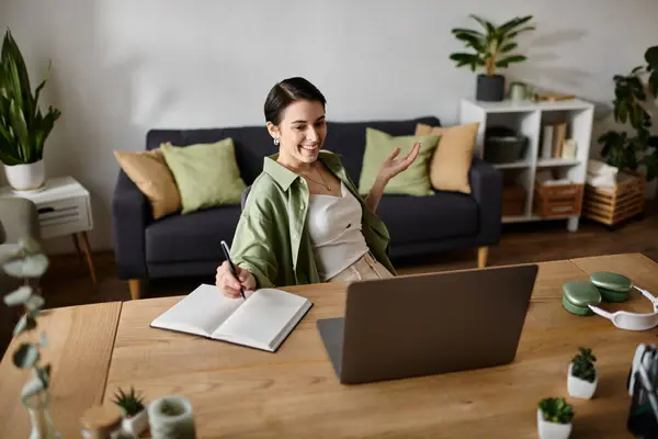 A woman smiles while working from home, sitting at a desk with a laptop and a notebook. — Stock Photo