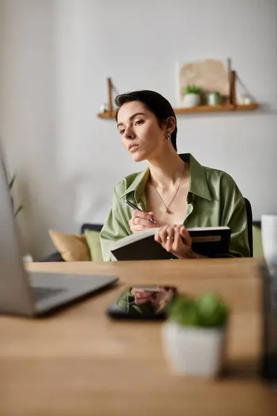 A woman sits at a desk in her home, working on her laptop and taking notes in a notebook. — Stock Photo