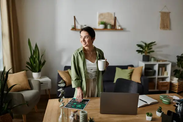 A woman stands in her home office, holding a cup of coffee and looking out the window. — Stock Photo