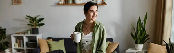 A woman in a green shirt smiles as she enjoys a cup of coffee in her home office. — Stock Photo