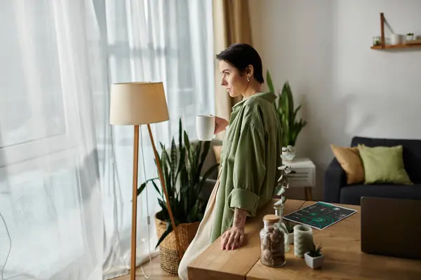 Uma mulher está em uma mesa em seu escritório em casa, segurando uma xícara de café e olhando pela janela. — Fotografia de Stock