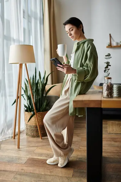 Woman in green shirt and beige pants leans on table with coffee and phone. — Stock Photo