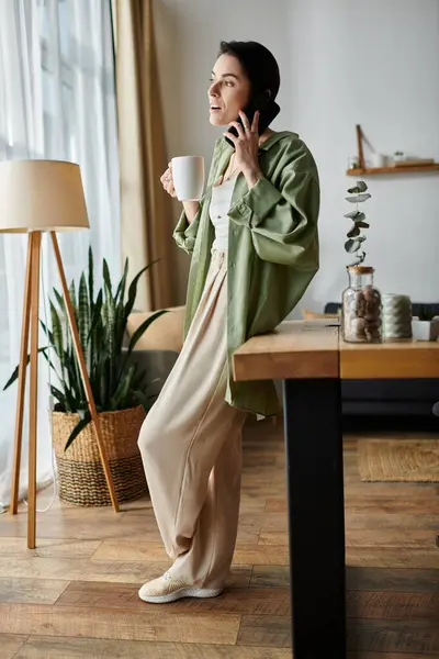 A woman in a green shirt stands by a window, talking on the phone while holding a cup of coffee. — Stock Photo