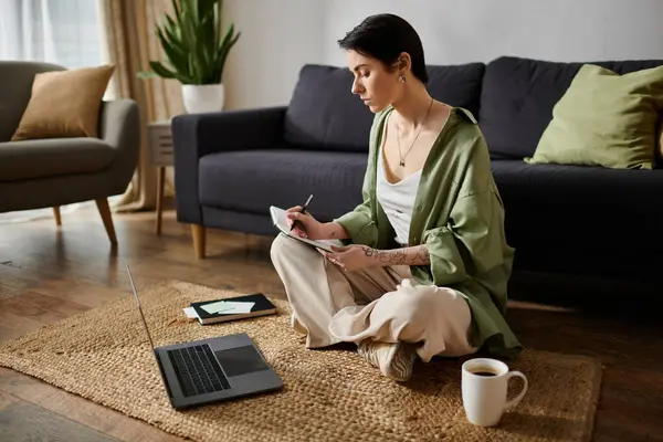 A woman sits on a rug in her living room, working on a laptop and taking notes in a notebook. — Stock Photo