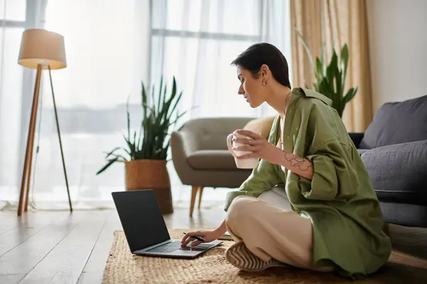 A woman sits on a rug in her living room, working on her laptop and enjoying a cup of coffee. — Stock Photo