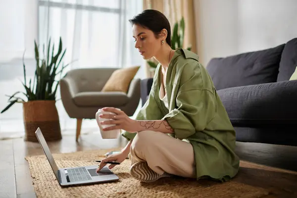 An attractive woman sits on the floor, working on her laptop with a cup of coffee in her hand. — Stock Photo