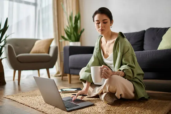 A woman sits on a rug in her living room, working on her laptop while enjoying a cup of coffee. — Stock Photo