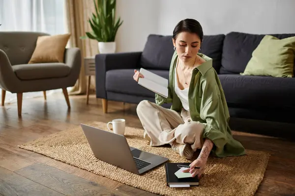 A woman sits on a rug in a living room, working on a laptop and taking notes. — Stock Photo