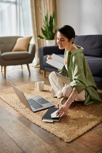 A woman sits on a rug in her home, working on a laptop and writing in a notebook. — Stock Photo