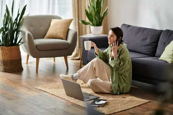 A woman sits on a rug with a laptop, a cup of coffee, and a phone, working remotely from her home. — Stock Photo