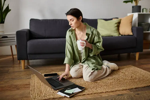 A woman sits on a rug in her home, working on her laptop and holding a mug. — Stock Photo