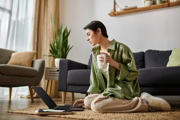 A woman sits on the floor in her living room, working on her laptop while enjoying a cup of coffee. — Stock Photo