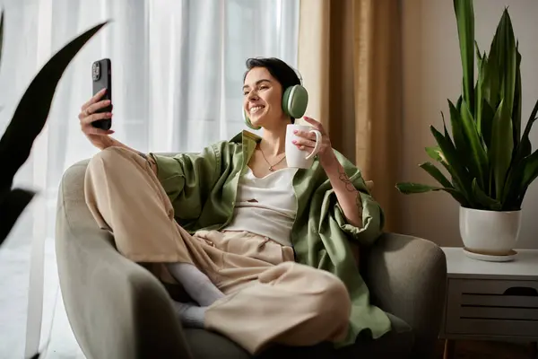 A woman sits in a chair at home, wearing headphones, holding a phone and drinking coffee. — Stock Photo