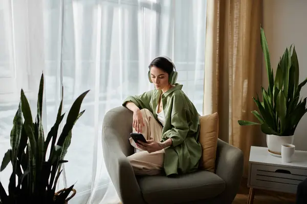 Eine Frau sitzt in einem bequemen Sessel am Fenster, trägt Kopfhörer und schaut auf ihr Handy.. — Stockfoto