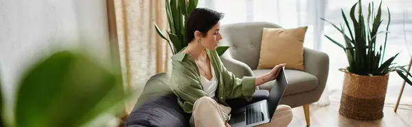 An attractive woman sits on a couch in a bright, home office space, working on her laptop. — Stock Photo