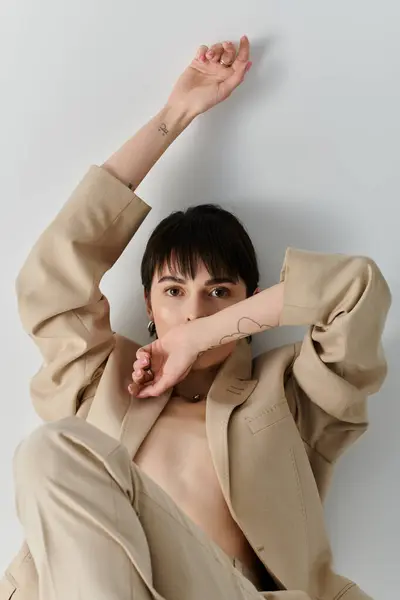 A woman wearing a beige blazer poses with one hand raised against a white wall. — Stock Photo