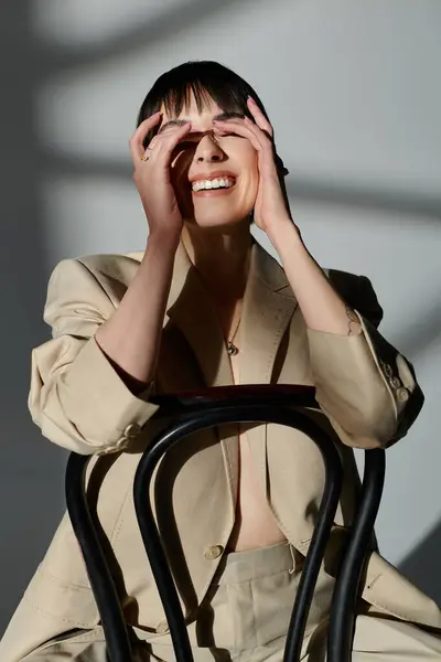 A woman laughs while sitting on a chair, her hands covering her eyes, in a bright studio setting. — Stock Photo