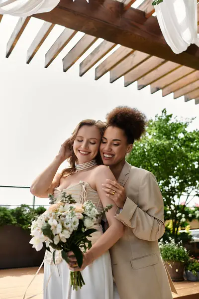 A lesbian couple embraces on their wedding day, standing under a wooden pergola. — Stock Photo
