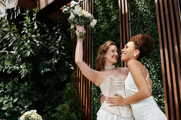Two brides share a loving embrace on their wedding day, surrounded by greenery. — Stock Photo
