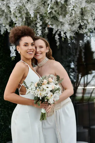 Two brides stand close, one holding a bouquet of white flowers, during their wedding ceremony. — Stock Photo