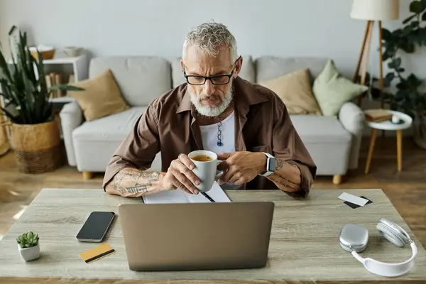 A mature gay man with grey hair and tattoos works from home, enjoying a cup of coffee. — Stock Photo