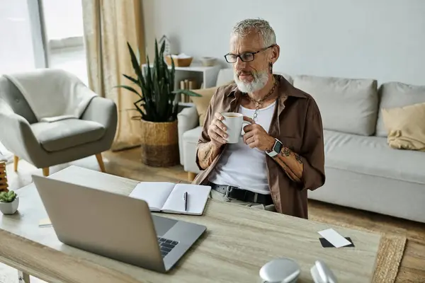 A mature gay man with tattoos and grey hair is working from home, drinking coffee and using a laptop. — Stock Photo