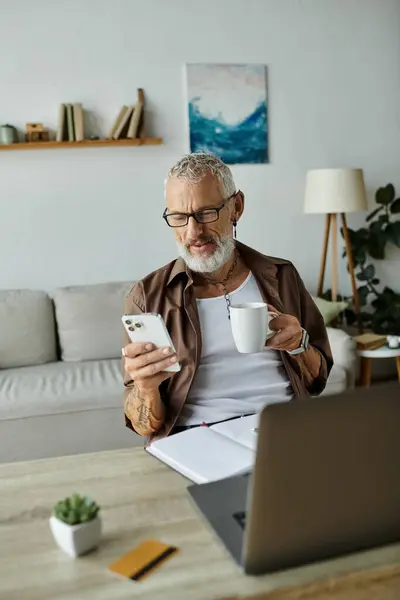 A tattooed, mature gay man with grey hair works remotely from home while holding a coffee mug and looking at his phone. — Stock Photo