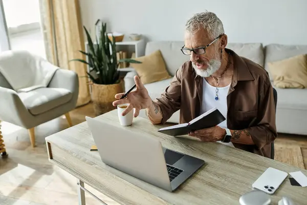 A mature gay man with tattoos and grey hair is working remotely at home, using a laptop and a notepad. — Stock Photo