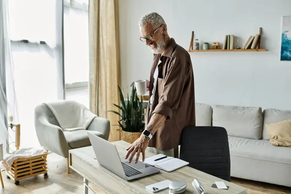 A mature gay man with tattoos and gray hair works remotely from his home office, holding a coffee mug and looking at his laptop. — Stock Photo