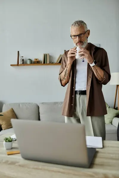 Ein reifer schwuler Mann mit Tätowierungen und grauen Haaren, der von zu Hause aus arbeitet und dabei eine Kaffeepause genießt. — Stockfoto