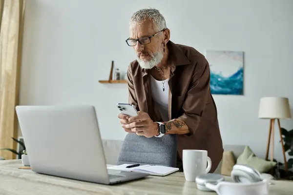 A mature gay man with tattoos and grey hair checks his phone while working from home. — Stock Photo