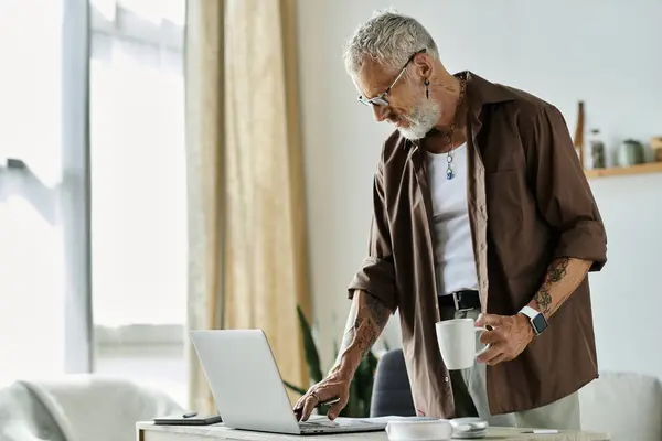 A mature, tattooed man with grey hair works on his laptop while holding a cup of coffee. — Stock Photo