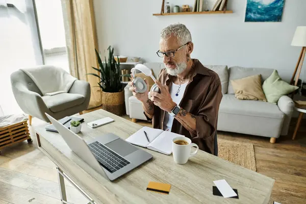 A mature gay man with tattoos and grey hair works remotely from home, holding headphones and looking at a laptop. — Stock Photo