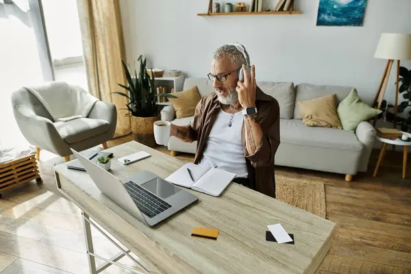 A tattooed man with grey hair works on a laptop at a desk, wearing headphones and holding a phone and coffee cup while working remotely. — Stock Photo
