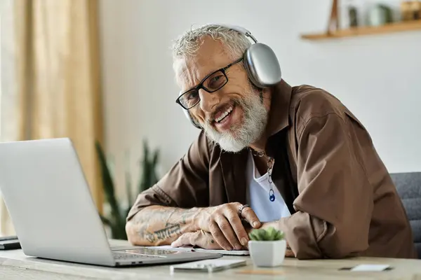 Ein reifer schwuler Mann mit Tätowierungen und grauen Haaren lächelt, während er von zu Hause aus auf seinem Laptop arbeitet. — Stockfoto