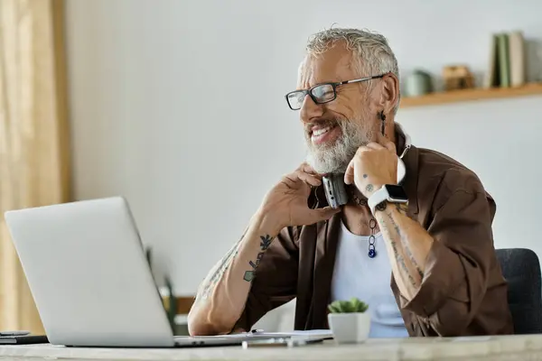 A mature gay man with tattoos and grey hair works remotely from home, smiling as he adjusts his headphones. — Stock Photo