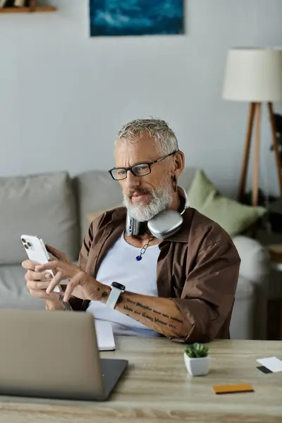 A mature man with tattoos and grey hair works remotely from his home. He is using a smartphone and a laptop while wearing headphones. — Stock Photo