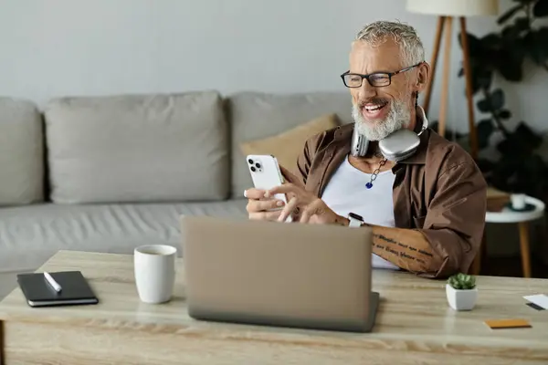 A mature gay man with tattoos and grey hair works remotely from home, smiling while looking at his phone. — Stock Photo