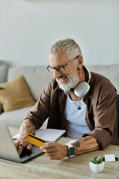 Um maduro gay homem com cinza cabelo e tatuagens funciona remotamente a partir de casa, usando um laptop e cartão de crédito, com um sorriso. — Fotografia de Stock