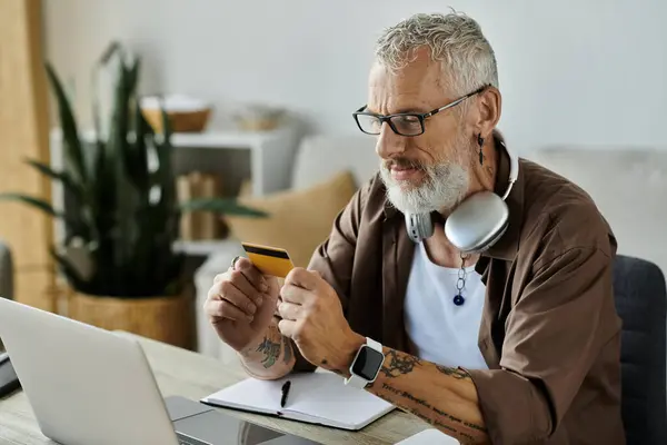 A mature gay man with tattoos and grey hair sits at his desk working remotely at home, holding a credit card and looking at his laptop. — Stock Photo