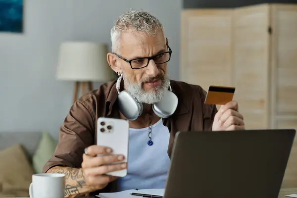 A mature gay man with tattoos and gray hair works from home, holding a credit card and smartphone. — Stock Photo