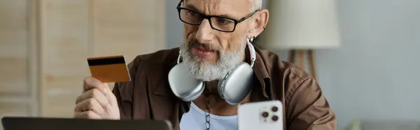 Un uomo maturo con capelli grigi, tatuaggi e cuffie lavora a distanza mentre tiene in mano una carta di credito. — Foto stock