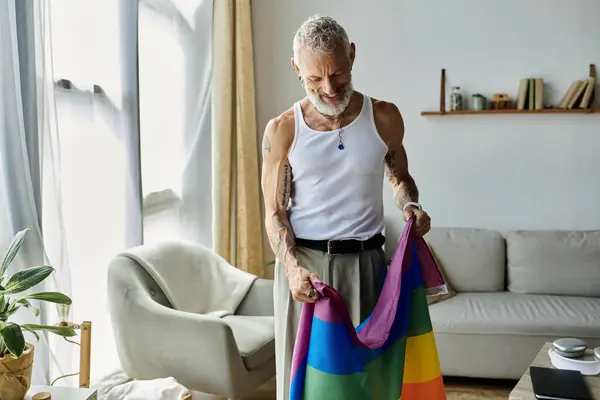 Um maduro gay homem com tatuagens e cinza cabelo detém um arco-íris orgulho bandeira no seu casa. — Fotografia de Stock