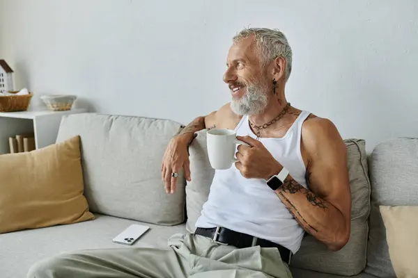 A mature gay man with tattoos and a grey beard relaxes on his couch at home, holding a mug of coffee and looking thoughtfully off-camera. — Stock Photo