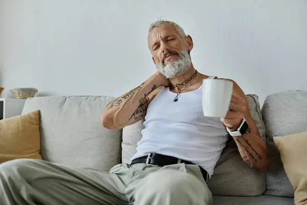 A mature gay man with tattoos and a grey beard relaxes on his couch at home, enjoying a moment of peace. — Stock Photo