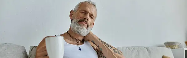 A mature gay man with tattoos and a grey beard relaxes on a couch at home, holding a cup of coffee. — Stock Photo