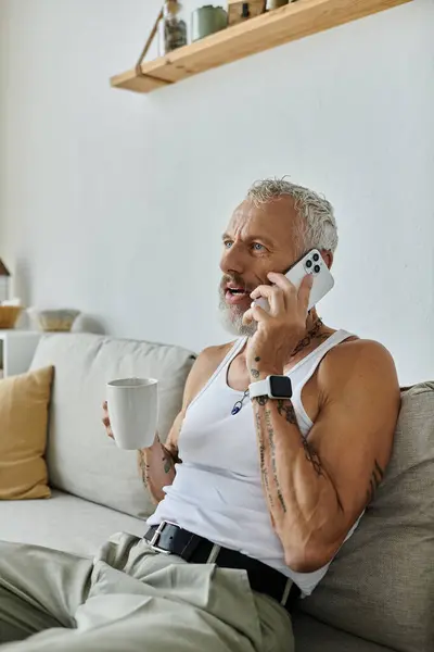 A mature gay man with tattoos and a grey beard sits on a couch at home, talking on his phone while holding a cup of coffee. — Stock Photo